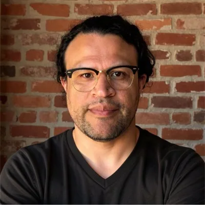 Walter Colindres, wearing glasses and a dark t-shirt, in front of a brick wall